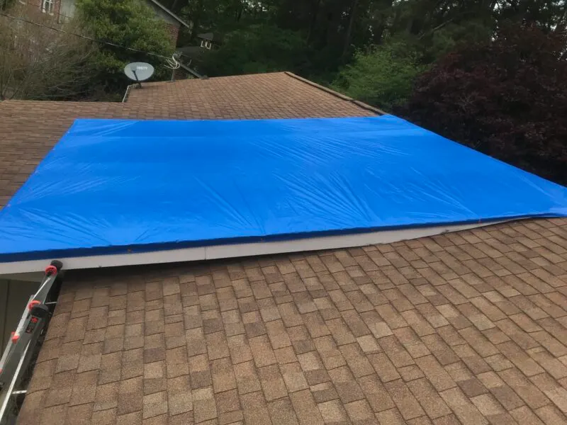 Blue emergency tarp secured over a damaged section of a shingle roof
